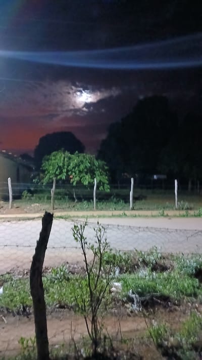 The image depicts an outdoor night scene at El Samán, Venezuela. In the night sky there is a full moon shining through the trees. There is a wooden fence with some greenery, dirt, grass, and a brick path. In the bottom right corner, there is the branch of a tree. There are also some houses in the background.