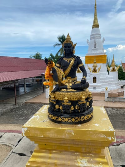 A black statue with gold accents depicting a seated figure wearing a crown and adorned with a gold sash and jewelry sits on a tiered, golden pedestal. The figure holds a yellow floral garland in its left hand. Behind the statue, a white stupa with a golden spire is visible, wrapped with a yellow sash. The stupa is surrounded by smaller golden spires. To the left of the statue, a building with a red corrugated metal roof and open-sided walkways with white railings is present. The ground is paved with hexagonal tiles. The scene is set outdoors under a partly cloudy sky. This image was taken in Baan Hua Thanon, Thailand.