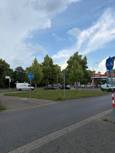 The image depicts a street scene in Singen (Hohentwiel), Germany, likely during the daytime, given the bright sky and clouds. The sky is a prominent feature, with a mix of white, wispy clouds and some larger, grayer formations against a blue backdrop. Lush green trees line the side of the road, suggesting a pleasant, possibly late spring or summer day.

The scene includes a road with a paved surface and a separate bicycle lane marked with a white bicycle symbol. There are also sidewalks and grass verges. Several vehicles are visible, including cars and a white van with "FS GROSSHAll" written on its side. The van appears to be a commercial vehicle. In the background, a gas station with "ESSO" signage is present, indicating a commercial area.

Two blue circular signs with a white bicycle symbol and a pedestrian are mounted on poles along the roadside, indicating designated paths for cyclists and pedestrians. These signs are a notable detail, highlighting the presence of infrastructure for non-motorized transport. The overall atmosphere seems calm and ordinary, depicting a typical urban or suburban street with traffic and amenities. The image is oriented vertically.