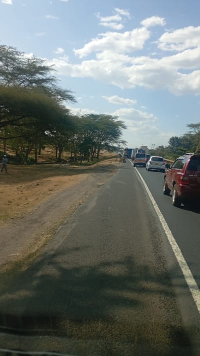 A slightly elevated view of a road with a double lane and a white line divider, traffic is visible. A red SUV sits on the right lane closest to the viewer. Across the road is a grassy verge leading to a tree line. Blue skies with puffy white clouds are overhead, the sun is shining casting shadows on the road. A person walks along the verge to the left of the road.