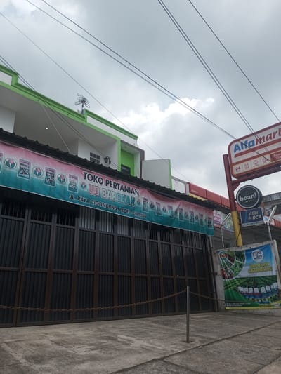 A street-level view captures a store front in what appears to be Indonesia, given the signage language. The primary storefront, "TOKO PERTANIAN UD BAROKAH", sells agricultural supplies. The building's exterior is predominantly white with green trim, featuring a horizontal banner above the entrance displaying the store's name and location: JL. RAYA CIGOMBONG, PACET-CIANJUR. The entrance is secured with a dark metal gate, slightly ajar.
Next to the store on the right is an "Alfamart", a convenience store chain. Additional signage advertises "bean! SPOT" and "ATM BCA". There is also a vertical banner for a product called "BAROKAH CIGOMBONG".
The scene is set against a cloudy sky, suggesting an overcast day. Utility wires are visible crisscrossing the sky. The overall environment implies a mix of commercial and potentially residential areas, given the presence of both agricultural supply and convenience stores. There are no people in the picture.