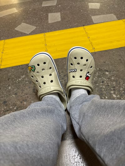 The image shows a pair of light beige Crocs with blue trim, adorned with two Jibbitz charms: one is an "I heart" symbol and the other is a palm tree with a yellow fruit. The Crocs are worn with light grey sweatpants and white socks. The scene is set on a speckled grey floor with a prominent, raised yellow line, possibly a tactile warning strip. The lighting suggests it is daytime, and the setting appears to be an indoor or covered area due to the smooth, manufactured flooring and the absence of natural outdoor elements. The "Crocs" logo is visible on both shoes.