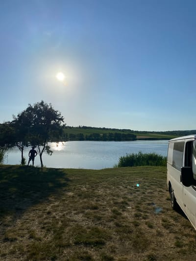 This image depicts a serene natural scene in Găleștii Noi, Moldova, under a clear blue sky. The sun is shining brightly, suggesting it is likely midday or late afternoon. The landscape is divided into a tranquil body of water on the left and a grassy field on the right. The water reflects the sky and the surrounding greenery, creating a shimmering effect. Along the edge of the water, a line of trees can be seen, adding depth to the scenery.
On the right side of the image, a white van is parked partially on the grassy terrain. The grass appears dry and somewhat sparse in places, with patches of longer green vegetation closer to the water's edge. The overall atmosphere is peaceful and idyllic, with a sense of quiet enjoyment of the outdoors.
In the foreground, silhouetted against the bright sun, a person stands near a tree by the water. The figure appears to be a man, standing casually and facing away from the camera, looking out at the water. The presence of the person adds a touch of human scale and suggests someone is enjoying the natural beauty of the location. The image conveys a sense of calm and relaxation. There is no visible text in the image.