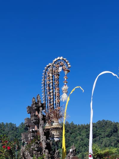 The image depicts a vibrant scene with a clear blue sky dominating the upper portion. In the foreground, a tall, ornate, tiered structure, possibly a temple decoration or a ceremonial arch, is adorned with intricate carvings and colorful elements. To the left of this structure, a part of a weathered, stone building with multiple levels and decorative carvings is visible, suggesting an ancient or traditional architectural style.
Flanking the central decoration are two large, flowing banners. A bright yellow banner curves gracefully to the left, while a stark white banner drapes to the right. In the background, a dense forest of lush green trees covers a hillside. The overall impression is one of cultural significance and natural beauty, possibly captured during a festival or a special occasion in a location like Baturiti, Indonesia, known for its temples and natural landscapes. Red flowers are also visible in the lower left corner of the image.