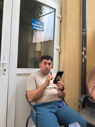 A medium shot shows a man seated outdoors, partially obscured by a window. He is wearing a light-colored t-shirt and blue pants. He holds a dark smartphone with a skeleton design in his right hand and a blue object, possibly a vape pen, in his left hand. To his left, a white doorframe with a handle and lock is visible. On the window, a blue sign reads "LOC PENTRU FUMAT", indicating a smoking area. Behind him, a dark pipe runs vertically along a textured yellow wall. The scene appears to be in Suceava, Romania, as suggested by the Romanian text on the sign. The man is looking slightly off-camera, towards the right.