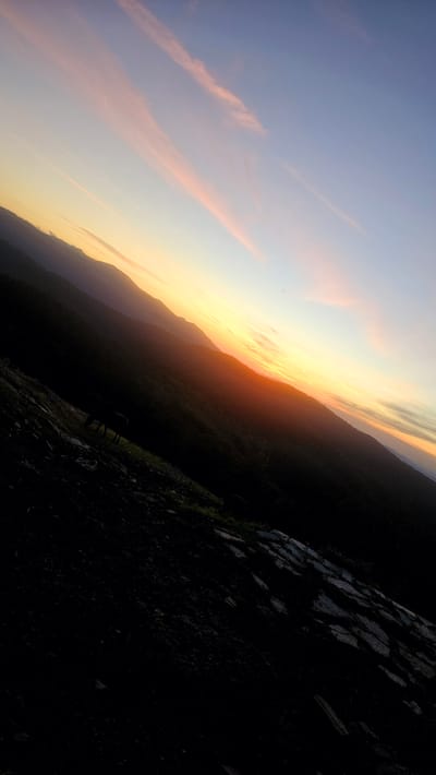 The image depicts a landscape scene, likely at dawn or dusk, characterized by a diagonal orientation. The sky transitions from a pale blue at the top to a warm yellow and orange near the horizon, with delicate pink clouds streaking across. Silhouetted against the bright sky are the layered shapes of distant mountains, their forms softened by atmospheric haze. In the foreground, the dark mass of a hillside slopes downwards, appearing rocky and uneven. A faint outline of what could be a horse or similar animal is visible grazing on a patch of green grass on the hillside. The overall impression is one of natural beauty and tranquility, with the low light casting deep shadows. The location in Simeonovets, Bulgaria, is not explicitly identifiable within the visual elements of the image itself.