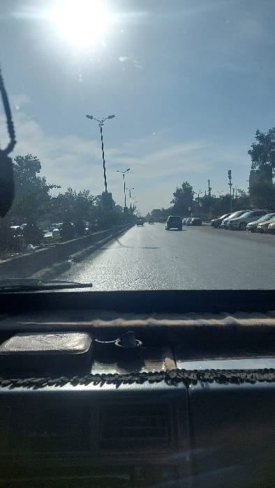 This image depicts a sunlit urban thoroughfare in Ash-Shaghour Municipality, Syria, captured from the interior of a vehicle looking forward through the windshield. The sky is bright blue with minimal cloud cover, and the sun, positioned in the upper left, casts significant glare and lens flare across the upper frame.

The central subject is a multi-lane asphalt road extending into the distance, with reflections of sunlight off its surface. A concrete median, topped with low barriers and green foliage, separates the lanes of traffic. Several vehicles are visible: two cars are traveling away from the camera in the middle lanes, while multiple other cars are parked parallel to the right-hand curb, facing the same direction as the parked vehicles.

Along the road and median, tall streetlights with distinctive three-lamp configurations are present. Trees and bushes line both sides of the road, with some buildings partially visible beyond the vegetation in the distance, particularly on the right.

In the immediate foreground, the dark dashboard of the originating vehicle is visible, featuring a textured trim piece with a patterned edge running along the base of the windshield. On the dashboard, a rectangular, metallic object is positioned on the left, and a dark, cylindrical container or holder is located centrally. A dark object, possibly a rearview mirror or hanging ornament, is partially visible on the upper left edge, suspended from the vehicle's interior. No individuals are clearly discernible.