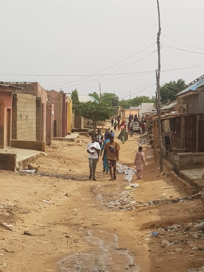 The image depicts a street in Dabawa, Nigeria. It is a dirt road with buildings on either side, some appearing to be unfinished or in a state of disrepair. There is a utility pole with visible wiring on the left. A group of people are walking along the road; their clothing is diverse. Litter and debris are scattered along the side of the road. A vehicle, potentially a small truck, is further down the road, surrounded by more people. The overall impression is one of an impoverished or underdeveloped area.