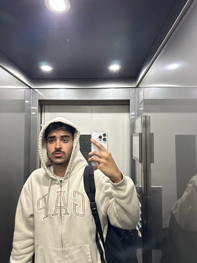 The image displays a single adult male taking a mirror selfie inside an enclosed space, likely an elevator. The male, with dark hair and a mustache, is shown from the upper torso, wearing a light beige zip-up hoodie featuring a cream-colored shearling-like lining in the hood. The word "GAP" is embroidered in large, outlined, light brown letters on the front of the hoodie. A dark-colored backpack is visible over his right shoulder. He holds a white smartphone with a multi-lens camera in his right hand, pointed towards the reflective surface to capture his image. The environment is characterized by silver-grey, highly reflective metallic or mirrored panels on the walls and ceiling. Two distinct circular recessed lights illuminate the ceiling. A portion of a silver-grey elevator door is visible on the left side, and a reflective panel with an indistinct control interface is present on the right. No identifiable architectural features, specific signage, or landmarks are visible within the image to confirm the precise location in Guimarães, Portugal.