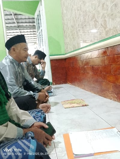 The scene takes place in a room with tiled floors and brick walls. The room appears to be part of a home or a small community building.  There are four men sitting on the floor. One man is wearing a blue shirt, black pants, and a black cap. He is seated at the front of the image, looking at his phone. The second man is wearing a grey shirt, black pants, and a black cap. He is seated behind the first man, looking down at a small book. The third man is wearing a green shirt, black pants, and a black cap. He is seated behind the second man, looking down at a small book. The fourth man, partially obscured by the first man, appears to be wearing a dark blue shirt. They are all sitting together, possibly engaging in conversation or a religious study session.  The room also contains two notebooks on the floor in front of them.  The location of this scene is Anjatan, Indonesia, at coordinates -6.419 latitude and 107.979 longitude.