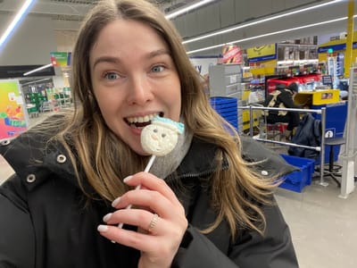 The image, originally oriented upside down, depicts a young woman in an indoor retail setting, possibly a supermarket or large store. She is looking directly at the camera with an open-mouthed, cheerful expression, revealing her teeth. Her light brown hair, which appears to have blonde highlights, falls around her face and shoulders. She is wearing a dark, possibly black, puffer jacket over a grey top. On her right ring finger, she wears a gold-colored ring with multiple thin bands. Her fingernails are painted a light, neutral color.
The woman is holding a white stick lollipop near her mouth, slightly to her left side. The candy portion of the lollipop is white and shaped like a round head, with a small blue detail at the bottom that resembles a scarf or a hat, giving it the appearance of a snowman or a cartoon character.
The background suggests a commercial environment. To her left, shelves are visible, holding various products and some blue bins or containers. The shelves appear to be well-stocked. To her right, more store shelves with colorful displays are present. A visible sign in the background, written in what appears to be Cyrillic script, clearly reads "СВОЙ MARKET" (SVOY MARKET), and below it, another word that looks like "СКИДКИ" (SKIDKI), which translates to "DISCOUNTS". The store has overhead fluorescent lighting, and the floor appears to be a light color. The background elements are slightly out of focus, drawing attention to the woman and the lollipop.