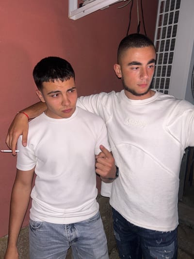 Two young men are lying on what appears to be an outdoor surface. The man on top wears a white sweatshirt with the text "Parental Philosophy" written on it, along with dark denim jeans. He also wears a watch on his left wrist and points with his right index finger. The man below wears a white waffle knit t-shirt and light blue jeans. He holds what looks like a cigarette in his left hand, while his right arm is around the first man's shoulder. The environment suggests an urban setting. There is no specific indication of Elhovo, Bulgaria. The background includes a red-painted wall, a white air conditioning unit, and other indistinct objects.
