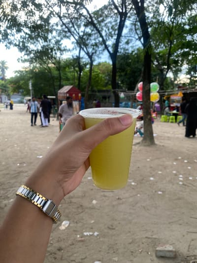 In Tarabo, Bangladesh, a person's left hand, adorned with a silver and gold watch, holds a transparent plastic cup filled with a pale yellow beverage. The cup is positioned in the foreground, partially obscuring the sandy ground dotted with white specks. In the background, a bustling outdoor environment unfolds. Several trees with lush green foliage stand tall, their branches reaching towards a bright sky. Beneath the trees, a few small stalls are visible, one with a red roof. People are scattered throughout the scene, some walking, others appearing to be interacting at the stalls. Colorful balloons are suspended in the air, adding a festive touch to the atmosphere. The ground appears to be a mixture of sand and dirt, with some scattered debris.