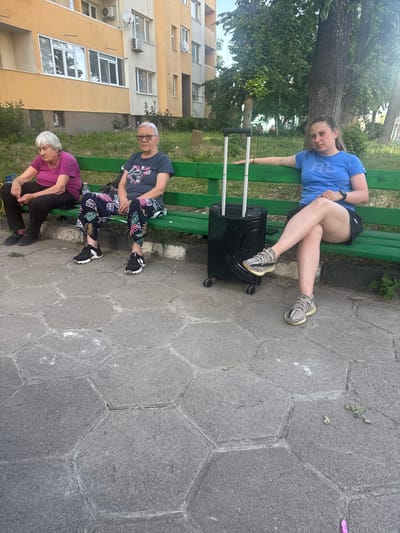 Three individuals are seated on a green park bench in front of an apartment building. The person on the far left, an elderly woman with gray hair, wears a purple t-shirt and dark pants. Next to her, another elderly woman with gray hair and glasses wears a dark patterned t-shirt and patterned pants. The person on the right, a younger woman with dark hair, wears a blue t-shirt and dark shorts. She is positioned next to a black rolling suitcase. The scene appears to be in a city park or residential area, with trees and foliage visible behind the bench. The ground is paved with hexagonal stone tiles. The lighting suggests it is daytime, possibly late afternoon due to the angle of the sun. This scene is located in Kyustendil, Bulgaria, as per the prompt.