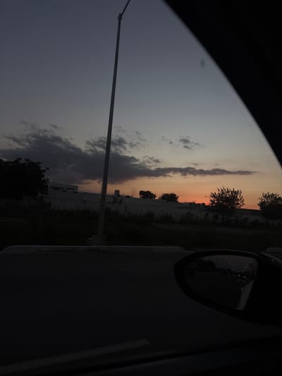 A view from inside a car captures a dusky sky transitioning to orange. A tall street lamp stands to the left against the gradient sky filled with clouds. A dark car door and side mirror dominate the right, reflecting lights. The landscape features a wall, trees, and building, all shrouded in darkness.