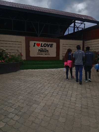 The image displays the exterior of the Riden Gate Resort, located in Limuru East ward, Limuru, Kenya. A large sign on a light-colored building facade centrally features "I ❤️ LOVE" in prominent letters, with "Riden Gate Resort" below it in a smaller, italicized font, and "LIMURU, KENYA" at the bottom. Below the sign is a strip of artificial green grass. Above the facade, a dark-colored, open-air structure with metal framework and a roof is visible against a grey, overcast sky.
A wide pathway paved with light-colored, scalloped interlocking concrete pavers occupies the foreground. Four individuals are walking away from the viewer on this path, towards the resort's entrance. From left to right: a person with long dark hair wearing a dark top and a bright pink skirt or shorts; a male in a grey t-shirt with "RIDEN..." visible on the back, and blue jeans; a male in a dark grey t-shirt with "RIDEN..." visible on the back, and dark pants; and a partially obscured fourth person on the far right, carrying a light blue bag. To the left of the pathway, a garden bed with green foliage and pink flowers is present.