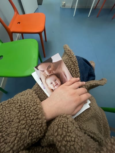 A person in a brown sherpa coat holds a photograph of a woman and baby with a cleft lip. A blue denim hat with sherpa trim sits on the arm of a lime green chair. An orange chair, a white counter, and wall-mounted coat hooks are in the background. The background is a pale blue wall with a gray floor.
