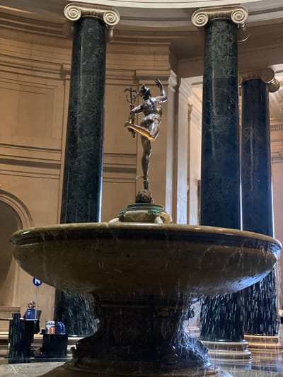 This is a medium-shot, eye-level, interior photograph of the Mercury Fountain at the National Gallery of Art in Washington, D.C. The bronze sculpture of the winged god Mercury stands atop a fountain that is actively spraying water. The fountain is surrounded by large, dark green marble columns with white Ionic capitals. In the background, beige walls with architectural detailing and other columns are visible. To the left of the fountain, a woman is seated at a desk with a blue sign and brochures, possibly an information or ticket booth. The scene is lit by artificial indoor lighting, casting highlights and shadows.