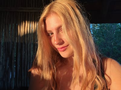 The image depicts a young woman with long, golden-blonde hair and light skin. She has brown eyes and a slight smile. The lighting is warm, possibly from the setting sun. She is wearing a delicate silver necklace with a small pendant. The background to her left consists of vertically arranged wooden slats, with shafts of light shining through. To her right, foliage is visible, suggesting an outdoor setting. The location is likely Khwai, Botswana, given the prompt's specification. The overall scene suggests a natural, possibly rural environment.