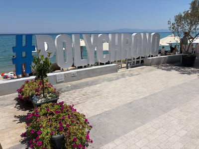 A bright, sunny day at a beachside location in Pallini Municipal Unit, Greece. The primary subject is a large, white, three-dimensional sign spelling out "POLYCHRONO", preceded by a blue hashtag symbol. The sign is positioned on a low concrete wall overlooking the turquoise sea. In the background, beach umbrellas, lounge chairs, and several people are visible, suggesting a relaxed atmosphere at a resort or beach club. A young man in a green t-shirt stands near a potted olive tree to the right of the sign, looking out towards the sea. In the foreground, two large planters overflow with vibrant purple petunias, one containing a small tree and white stones. The ground is paved with light-colored bricks. The clear blue sky and calm water indicate pleasant weather. A few white signs are affixed to the concrete wall behind the planters and sign. One sign has the text "IAS" and "AIAS".