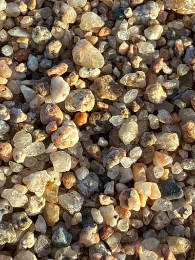 This media file shows a close-up, overhead view of a collection of small, irregularly shaped pebbles and sand grains. The subjects are the pebbles and sand, which vary in size, color, and texture. Colors range from white, clear, and light yellow to tan, orange, brown, gray, and black. Some pebbles are translucent, allowing light to pass through them, while others are opaque and speckled. The texture appears rough and granular overall, with some individual stones showing smoother facets.
There is no discernible activity or event taking place; the media file is a still image of the static collection of stones.
Notable details include the lighting, which suggests a bright, sunny day, casting distinct shadows among the pebbles and highlighting their surfaces. The light appears to be coming from the upper right. The scene is likely outdoors, given the natural materials and the quality of light. There are no explicit location cues other than the provided context of Blanes, Spain, which might suggest a beach or riverbed environment, though this cannot be confirmed solely from the image. No people are visible, and no emotions are conveyed.
There is no visible text in the media file. The overall impression is a detailed, textural study of natural mineral aggregation.