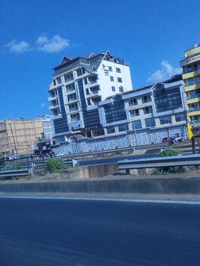 A tall, modern hotel complex stands beside a road. The building features a mix of white sections with many windows and a taller section with balconies. The word "HOTEL" is visible above the entrance. A few other buildings are seen in the background, and there's a guardrail separating the road from the hotel. The sky is bright blue with a few clouds.