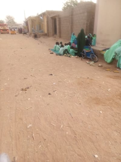 A group of children in green uniforms are seated on mats along a dusty street. An adult figure in a long green robe and veil stands beside them. A blue wheelchair is parked near the group. Various pairs of shoes and sandals are scattered around the children. In the background, a worn building facade and distant vehicles can be seen. The ground is littered with debris.