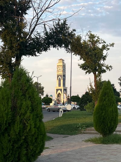 A clock tower is visible in the distance, framed by the branches of two trees and two green cypress bushes in the foreground. The clock tower is tall and rectangular, with a white and blue facade and a pointed roof. It has a clock face on each of its four sides. The clock tower is located in the city of Taraz, Kazakhstan, and it is the administrative building of the city. In the middle ground, cars are parked on the side of the road. The ground in the foreground is paved with cobblestones, and there is a lawn area with grass and some small bushes. The sky is partly cloudy and appears to be either early morning or late afternoon. The scene is peaceful and serene, with no people or activity visible in the immediate foreground.