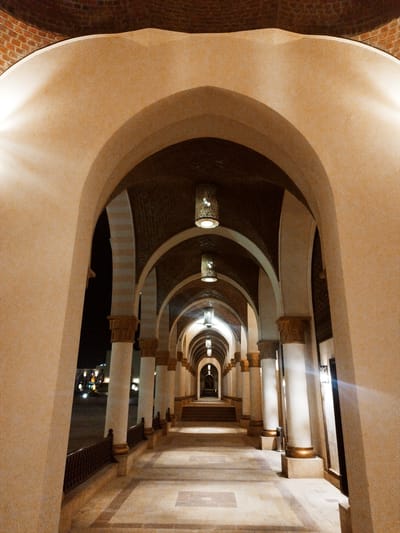 The image shows an illuminated arched corridor in an architectural structure. The structure appears to be located outdoors, as indicated by exterior lights visible in the background. The corridor consists of multiple arches supported by pillars, with decorative lighting fixtures hanging from the ceiling. The corridor leads to a dark area. The floor is paved with tiles. The location is labeled as Unknown, Egypt.