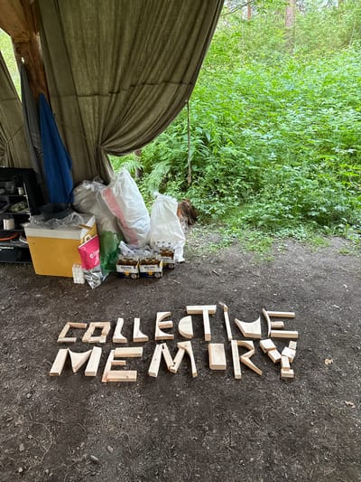 In an outdoor setting, likely in the city of Cheposh, Russia, an arrangement of wooden blocks spells out "COLLECTIVE MEMORY" on a dirt ground. To the left, a makeshift shelter is visible, draped with a green canvas. Several white and pink bags, a yellow cooler, a green plastic bag, and six bottles of Corona Extra beer are placed near the shelter. The background is filled with lush green foliage and trees.