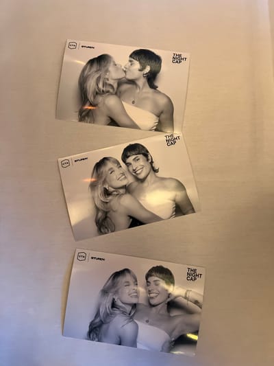 The image shows three identical promotional postcards for "The Night Cap", a production of Uta and Sturdy, set against a white textured surface, possibly a tablecloth or bedsheet. Each card features a black and white image of two women in a romantic embrace. The name of the production, "The Night Cap", is printed vertically near the top left of each card, and the Uta and Sturdy logos are vertically aligned near the bottom left corner. The postcards are laid side by side, with slight variations in angle and lighting, suggesting they are photographed in a casual setting. The image does not show the city of Indian Wells, United States of America.