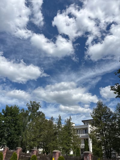 The image depicts a scenic view of a blue sky with scattered white clouds. The clouds are a mix of wispy cirrus formations and larger cumulus clouds, suggesting a dynamic weather pattern. The sky takes up a significant portion of the frame, creating a sense of vastness.
In the lower right portion of the image, trees with lush green foliage are visible, bordering a wooden fence. Behind the trees, a modern building with white walls and multiple windows can be seen. The architecture suggests a residential or possibly commercial property. The trees are casting shadows, and the sunlight filtering through the leaves indicates it is likely daytime, possibly late morning or early afternoon.
The overall mood of the image is peaceful and natural, with the bright sky and greenery creating a pleasant atmosphere. The location context provided is "Парэчча, Belarus," which places the scene in a specific geographical area. There is no visible text in the image. The composition is vertical, with the sky dominating the upper left and the trees and building occupying the right side.
