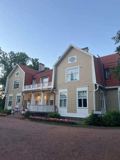 The image shows the exterior of a two-story, light yellow wooden house with a red tiled roof. The house features multiple windows, a covered porch with white railings and pillars, and a set of stone stairs leading up to the porch. Flower beds with pink and purple flowers are visible in front of the house, and a gravel path runs in the foreground. A ladder is propped against the side of the house, leaning against the roof. A sign with text in Finnish, "Päärakennus Håkansböle kartanomuseo", is visible near a bush. The house is surrounded by trees and the sky is clear and blue. This is the main building of the Håkansböle Manor Museum in Vantaa, Finland. No people or animals are present in the image.