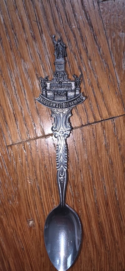 A detailed, ornate silver spoon lies on a wooden surface. The spoon's handle is decorated with intricate scrollwork and culminates in a detailed depiction of the Niederwald Monument. This monument features a prominent statue of Germania at the top, with smaller statues and architectural elements below, including the inscription "NIEDERWALD DENKMAL" across a banner. The wooden surface exhibits a prominent grain pattern and appears to be a table or floor. The image is a top-down, close-up shot, with the spoon centrally placed. The Niederwald Monument is located in Germany, not Marum, Netherlands.