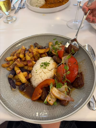 A top-down view shows a meal served in a bowl on a table, likely in a restaurant. The bowl contains a mound of white rice, surrounded by fried potatoes with a purple hue, pieces of beef, sliced red tomatoes, sliced onions, and garnish. A metallic spoon is visible near the top left of the bowl, and another dish is partially visible to the left, containing noodles and rice. Cutlery is seen on the table near the bottom left. The setting is likely indoors, possibly in a restaurant. While the specific location in Coral Gables, United States of America, is not definitively ascertainable from the image alone, the style of the dish suggests a restaurant serving Peruvian cuisine.