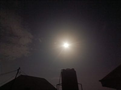 The media file depicts a nighttime scene in Galadimawa Village, Nigeria, dominated by a bright moon in a cloudy sky. The moon is the central subject, casting a strong glow and creating visible rays, indicating it is either full or near-full. Scattered clouds partially obscure the moon, adding texture to the dark sky.
In the foreground, silhouetted against the moonlit sky, are elements of the village environment. A prominent object is a tall, cylindrical water tank supported by a metal frame, likely a common sight for water storage in the area. To the left, the silhouette of a roofline is visible, along with utility poles and wires, suggesting the presence of electricity. On the right, another roofline is partially visible, also silhouetted against the sky.
The overall impression is one of a quiet night. The illumination from the moon suggests it is nighttime, and the clear visibility of the moon despite some cloud cover implies fair weather, though the clouds indicate some atmospheric moisture. No people are directly visible in the frame. The scene is likely an outdoor view from a residential area within Galadimawa Village, capturing a moment of nocturnal tranquility illuminated by the moon. There is no visible text in the media file.