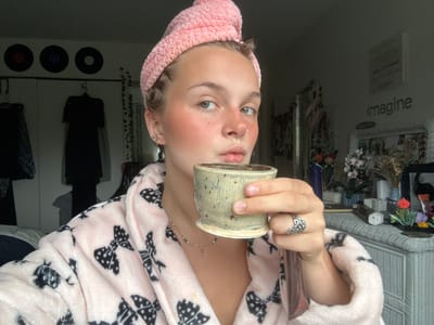 The image features a young Caucasian female indoors, likely in a bedroom. She is wearing a pink terry cloth hair wrap and a light pink plush robe adorned with black polka-dotted bow patterns. Her face shows light freckles on her nose and cheeks, pink blush, a silver stud earring in her left earlobe, and a silver hoop nose ring on her left nostril. She holds a speckled, rustic light-colored ceramic mug in her right hand, raised towards her mouth, and looks directly at the camera. A silver necklace with a small green or gold pendant is visible around her neck, and a silver ring with an intricate design is on her right ring finger.
The background on the left wall displays three dark vinyl records mounted above a clothes rack holding dark garments and white closet doors. Artificial ivy hangs down the wall. On the right, a white wicker-style dresser or cabinet is visible, topped with various decorative items including dried and artificial flowers, small vases, and cosmetic brushes. The word "imagine" is prominently displayed in gray lettering on the wall above the dresser, and a smaller, partially visible sign above it reads "MAKE A DIFFERENCE." The lighting appears to be natural, coming from the right side of the frame.
There are no discernible visual cues within the image to confirm the specified location as Whistler, Canada.