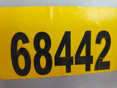 A close-up, eye-level, medium shot shows a bright yellow rectangular sticker with the number "68442" printed in bold black numerals. The sticker is horizontally oriented and appears to be affixed to a textured gray surface, possibly a wall or piece of equipment. The lighting is even, and there are no people or other distinguishing objects in the frame. The background is out of focus, revealing only the gray texture. This image contains no information about Tecnimont camp or Russia.