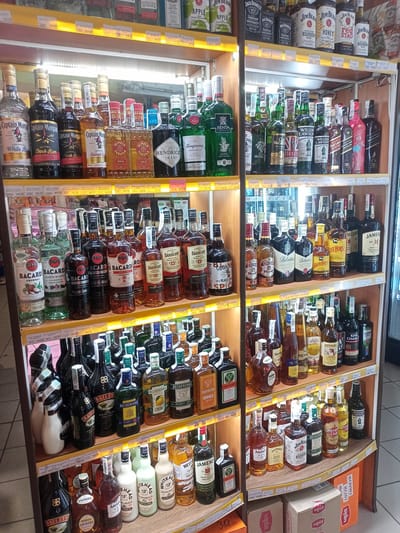 The image depicts a retail shelf displaying a variety of alcoholic beverages in a store. The products are organized on wooden shelves, with some items appearing to be more prominently displayed than others. Brands visible include Captain Morgan, Olmeca, Hendrick's Gin, Tanqueray, Bacardi, Oakheart, Jameson, Chivas Regal, Ballantine's, White Horse, Jim Beam, Jägermeister, and various other spirits and liqueurs. The lighting suggests an indoor environment, likely a liquor store or a section of a supermarket. There are price tags visible below some of the shelves. The background is blurred but appears to be additional shelving and possibly refrigerators. There are no people visible in the frame. The presence of product labels and the arrangement suggest a commercial setting for sales. Based on the provided context that the image is from Bilhorod-Dnistrovskyi, Ukraine, this scene is likely a liquor store or a beverage aisle within a supermarket in that city.