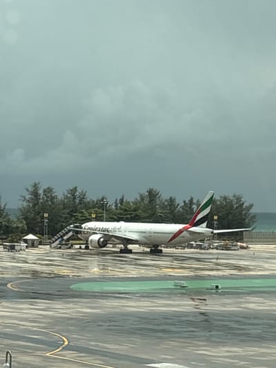 An Emirates airplane is parked on a tarmac in Mai Khao, Thailand, under a cloudy sky. The plane is white with red and green accents on its tail fin, and the Emirates logo is visible on its side. A set of stairs is positioned near the front of the plane, connecting it to the ground. Several vehicles, including baggage carts and a small white structure, are also present on the tarmac. In the background, there is a line of trees and the ocean. The tarmac appears wet, suggesting recent rain. The scene is captured from a slightly elevated perspective, possibly from within an airport terminal.