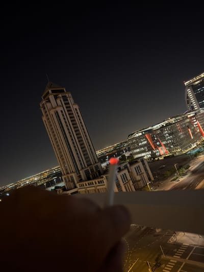 The image captures a nighttime cityscape from a high vantage point, likely a balcony or window of a tall building. The scene is dominated by modern, illuminated architecture under a dark, clear sky.
In the immediate foreground, to the right, a human hand is visible, partially obscured by a light-colored railing or frame. The hand holds a slender, pen-like object with a glowing red tip, which is directed towards the illuminated urban sprawl.
The midground and background feature several contemporary high-rise buildings. One prominent, tall rectangular building stands out with numerous illuminated windows, showcasing a sleek, modern design. Further into the city, another complex of buildings is visible, characterized by intricate facades and striking red, triangular architectural elements that glow brightly against the night.
Below the buildings, a network of well-lit roads is visible, featuring clear lane markings, pedestrian crossings, and streetlights. These roads weave through the urban landscape, connecting various structures. The city is extensively lit, with countless artificial lights from buildings and streets creating a vibrant and expansive urban glow that stretches into the distance. The overall impression is one of a bustling, modern city active after dark. No discernible text is visible anywhere in the image.