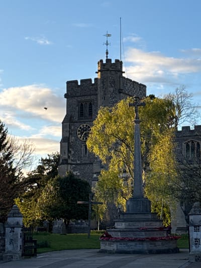 The image shows a vertical view of St Peter and St Paul Church in Tring, a town within the borough of Dacorum in Hertfordshire, United Kingdom. The stone church features a prominent clock face, a weather vane atop its tower, and a visible spire. A stone war memorial stands in the foreground, with red poppy wreaths affixed. The surrounding environment includes trees with green foliage under a partly cloudy blue sky. A small bird is visible in the sky near the clock.