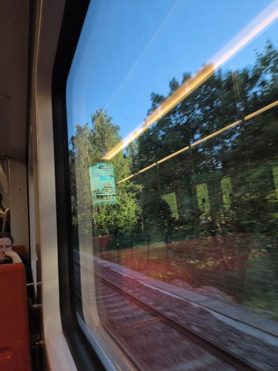 The image is a view from inside a train carriage in Helsinki, Finland, looking out the window. The window reflects the interior of the train, including a person seated in a red train seat. Outside the window, trees and a clear blue sky are visible, with train tracks in the foreground. The train appears to be moving, as indicated by the motion blur of the trees and the reflection. A blue poster is affixed to the inside of the window. The lighting suggests it is late afternoon or early evening.