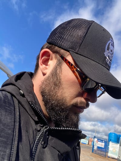 This is a close-up, eye-level, outdoor, medium-shot photograph of a man wearing a black mesh trucker hat, sunglasses with tortoiseshell frames, and a black hooded jacket. He has a beard and is looking down and to his right. The background consists of a blue sky with scattered clouds, industrial shipping containers, and a blue gate with signage. The location is identified as Koskolovo, Russia. The man's right ear is visible, and his black jacket has a prominent zipper closure. The lighting suggests it is daytime.