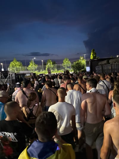 This image captures a nighttime gathering of people, likely at an outdoor event. The sky is dark with some clouds, suggesting it is evening or night. The scene is illuminated by artificial lights, some of which are visible as bright points in the sky and along a structure in the background. There are trees with green foliage visible, indicating an outdoor setting.

The subjects are predominantly people, many of whom are facing away from the camera or have their heads down, suggesting they are watching something in front of them. Some individuals are wearing t-shirts, and others are shirtless. The overall atmosphere appears to be one of a crowd at a concert, festival, or sporting event. There is no visible text in the image. The presence of a blue and yellow bag or flag could be a contextual detail, but its specific meaning is not discernible from the image alone.