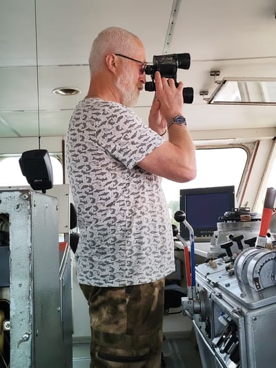 An older man with a grey beard and glasses is standing in what appears to be the bridge of a vessel, possibly a riverboat given the context of Yaroslavl, Russia, a city situated on the Volga River. He is holding a black monocular to his eyes, looking out towards the unseen exterior. He is wearing a white short-sleeved shirt with a pattern of grey sharks and camouflage-patterned pants. On his left wrist, he wears a silver watch. In the foreground, to the left of the man, is a black microphone attached to a cable. To the right of the man, a control panel with various levers and dials is visible, including a computer monitor with a dark screen. The environment suggests an operational setting, likely a control room or bridge of a boat, where navigation or monitoring activities are taking place.