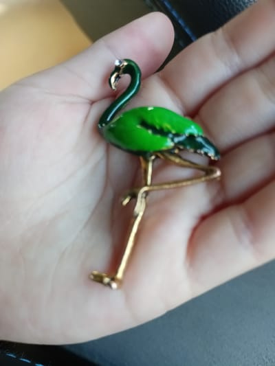 The image shows a green flamingo-shaped brooch resting on a person's hand. The brooch has gold-colored legs and beak, and a small gem on its head. The background is blurred, showing a beige and dark gray surface. The image does not provide any information about the location of the city of Ankara, Turkey.