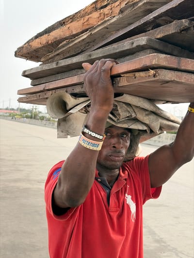 A man in a red Ralph Lauren polo carries a stack of weathered wooden planks on his head. The load is balanced with the help of a beige cloth underneath. He wears two wristbands on his right arm, one reading "BEYOND," the other reading "EXPECTATIONS." He gazes directly at the camera with a weary expression. The background shows a hazy cityscape.