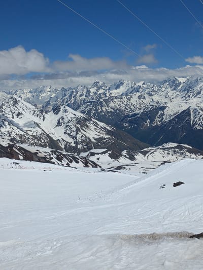 The image depicts a vast, snow-covered mountain range under a bright blue sky with scattered clouds. Several thin, white cables are stretched across the upper portion of the frame, disappearing into the distance. The mountains themselves are rugged, with sharp peaks and deep valleys, and are partially covered in snow, with darker rock faces exposed in places. In the foreground, a wide expanse of snow slopes gently downwards, marked with tracks indicating recent activity, possibly from skiing. There are no people visible in the image. The scene is characteristic of a high-altitude, mountainous environment, likely a resort area, and the presence of cables suggests a ski lift or similar infrastructure. Given the context of Terskol, Russia, this scene is consistent with the natural landscape surrounding the town, known for its proximity to Mount Elbrus, a prominent peak in the Caucasus Mountains.
