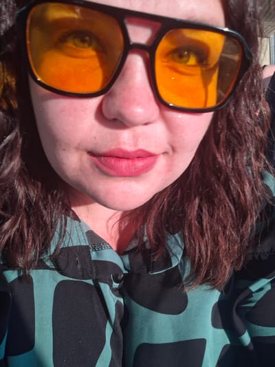 A close-up photograph depicts a person lying down with their head turned to the side. The person has fair skin, pink lips, and brown curly hair. They are wearing sunglasses with orange lenses and black frames. A green and black patterned shirt is visible on the left side of the image. The background is out of focus. The location is not discernible from the image, so it is not possible to determine if it is in Kamennyi Kliuch, Russia.
